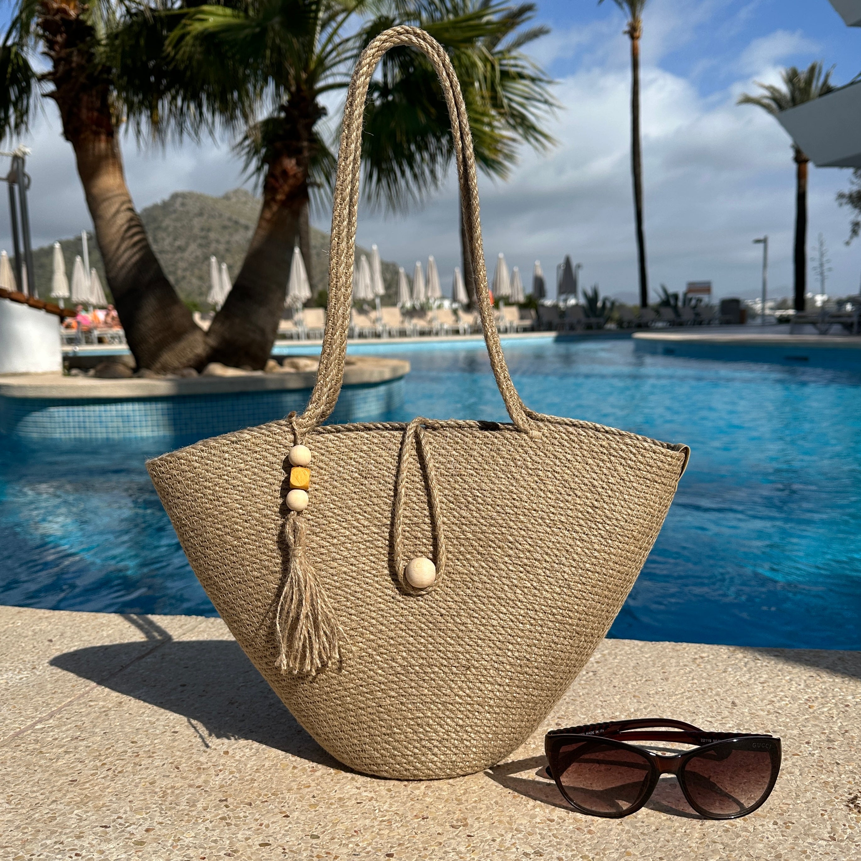 Håndlavet taske af naturlig jute med lange hanke, træknaplukning og dekorativ kvast med trædetaljer. Et stilfuldt sommeraccessory.