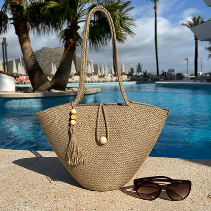 Håndlavet taske af naturlig jute med lange hanke, træknaplukning og dekorativ kvast med trædetaljer. Et stilfuldt sommeraccessory.
